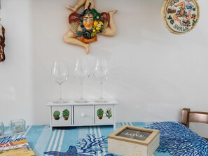 A room with a white cabinet holding three wine glasses, a small table with a blue tablecloth, and a decorative plate with 'Sicilia' on the wall. - Belvilla by OYO Bedda Sicilia Mia (Palermo)