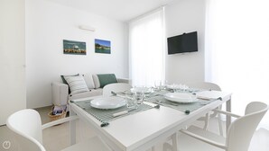 A modern, minimalist dining area with a white table set for four, surrounded by white chairs. There is a sofa with striped cushions in the background, and two framed pictures on the wall. - Rivazzurra Homes - 57 (Termoli)