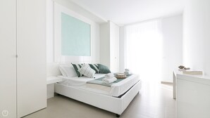 A modern, minimalist bedroom with a white bed, green and white striped pillows, a small bedside table with a book and a decorative item, and a large window with sheer curtains. - Rivazzurra Homes - 57 (Termoli)
