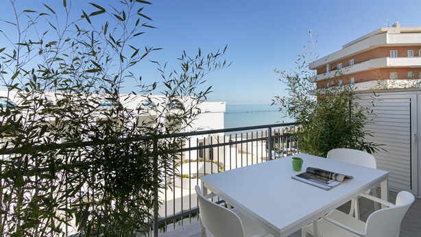 A balcony with a white table and chairs, overlooking a beach and a building. - Rivazzurra Homes - 57 (Termoli)