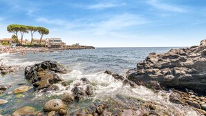 A rocky shoreline with clear water, a beach with people, and buildings in the background. - Holiday Home in Santa Tecla Near Etna Beach (Acireale)
