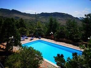Ein rechteckiger Pool mit Steinrand, umgeben von Liegestühlen und Sonnenschirmen, vor Hügeln und Bäumen.
