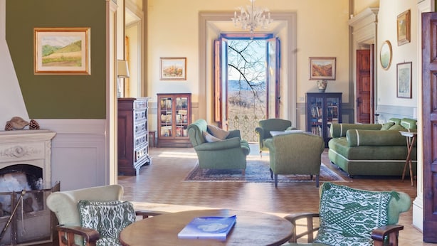 Un salon spacieux avec une cheminée, des fauteuils verts, une table basse en bois et une vue dégagée sur l’extérieur grâce à une grande fenêtre.