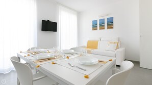 Un comedor moderno y bien iluminado con una mesa blanca dispuesta para cuatro personas, un televisor de pantalla plana en la pared y un sofá con cojines al fondo.