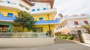 A multi-story residential building with a white facade, yellow and blue accents, and a white picket fence in the foreground. - Belvilla by OYO Appartamento Spinesante (Barcellona Pozzo di Gotto)