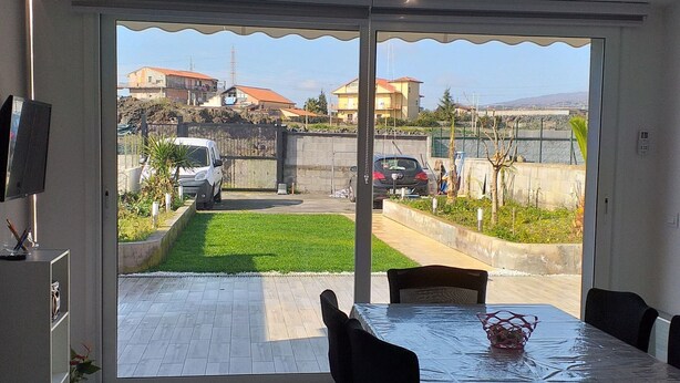 A dining area with a table and chairs, a TV on the wall, and a view of a residential area with houses and parked cars. - Belvilla by OYO La Casa di Giada (Belpasso)
