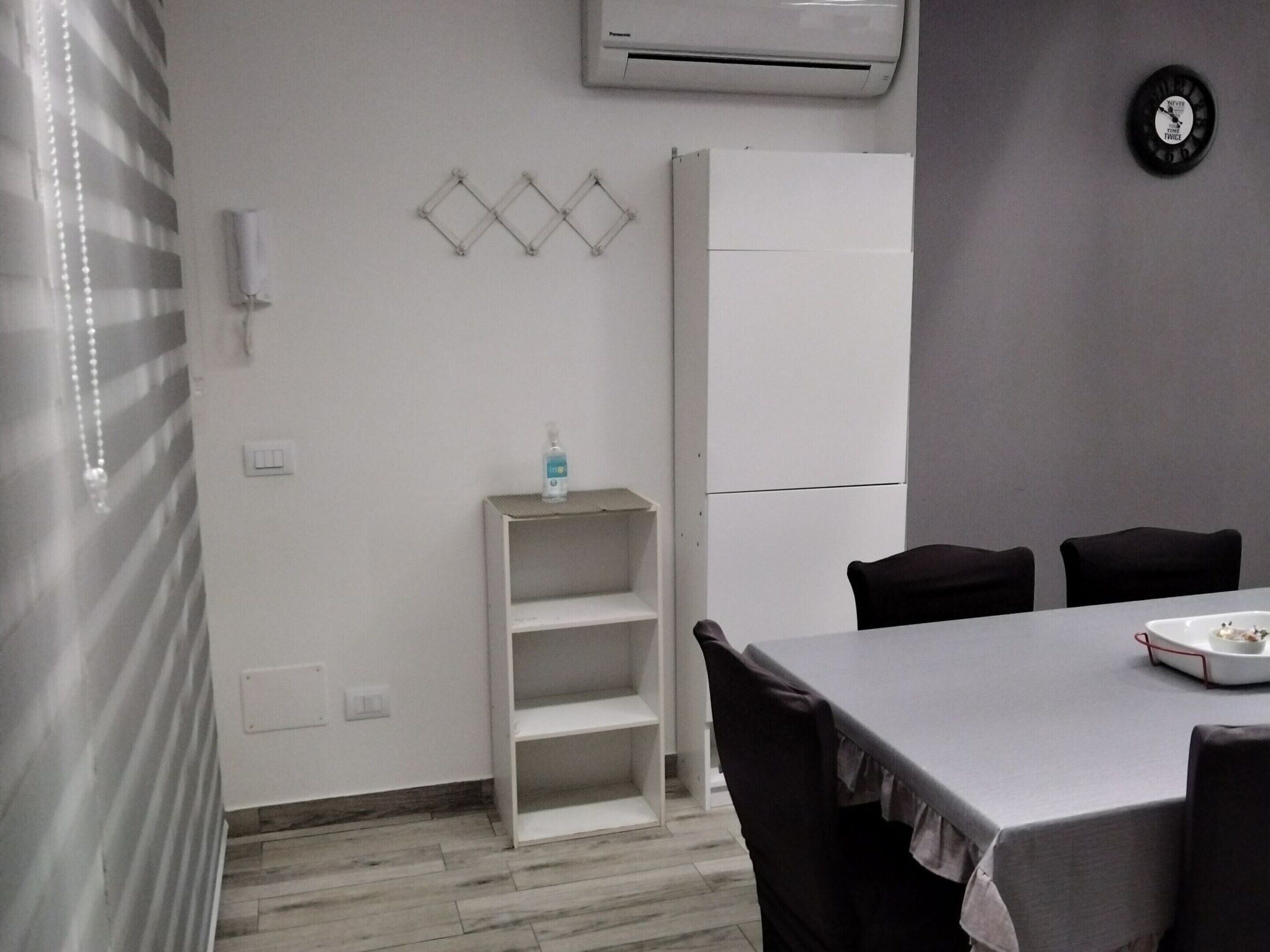 A small, modern kitchen with a white fridge, a white shelf unit, and a dining table with chairs.