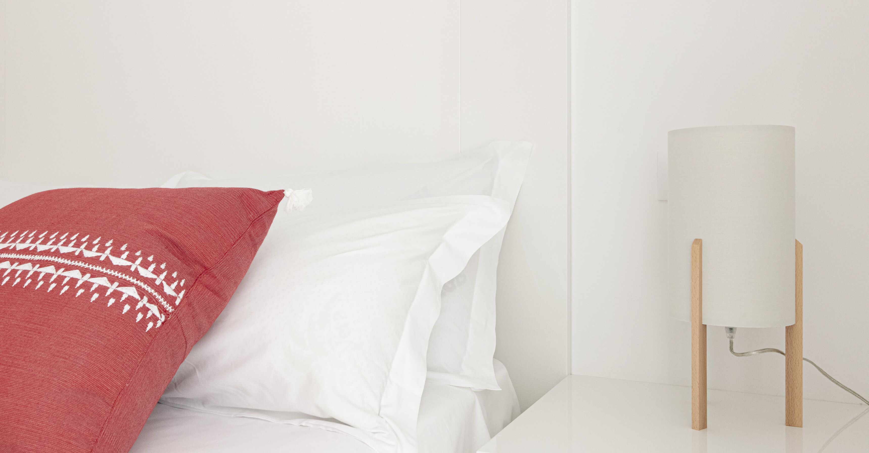 A bed with white linens and a red blanket with a white pattern, a white wall, and a white table with a lamp on it.