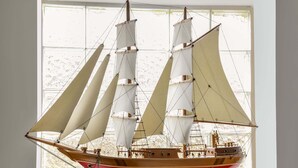 Um modelo de navio exibido em ambiente interno, suspenso por uma corrente, com uma janela ao fundo.
