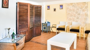 A living room with a glass coffee table, wicker sofa, and a white table. There are two chairs, a blue cabinet, and a stone wall. - Alluring Holiday Home in Syros With Barbecue (Syros)
