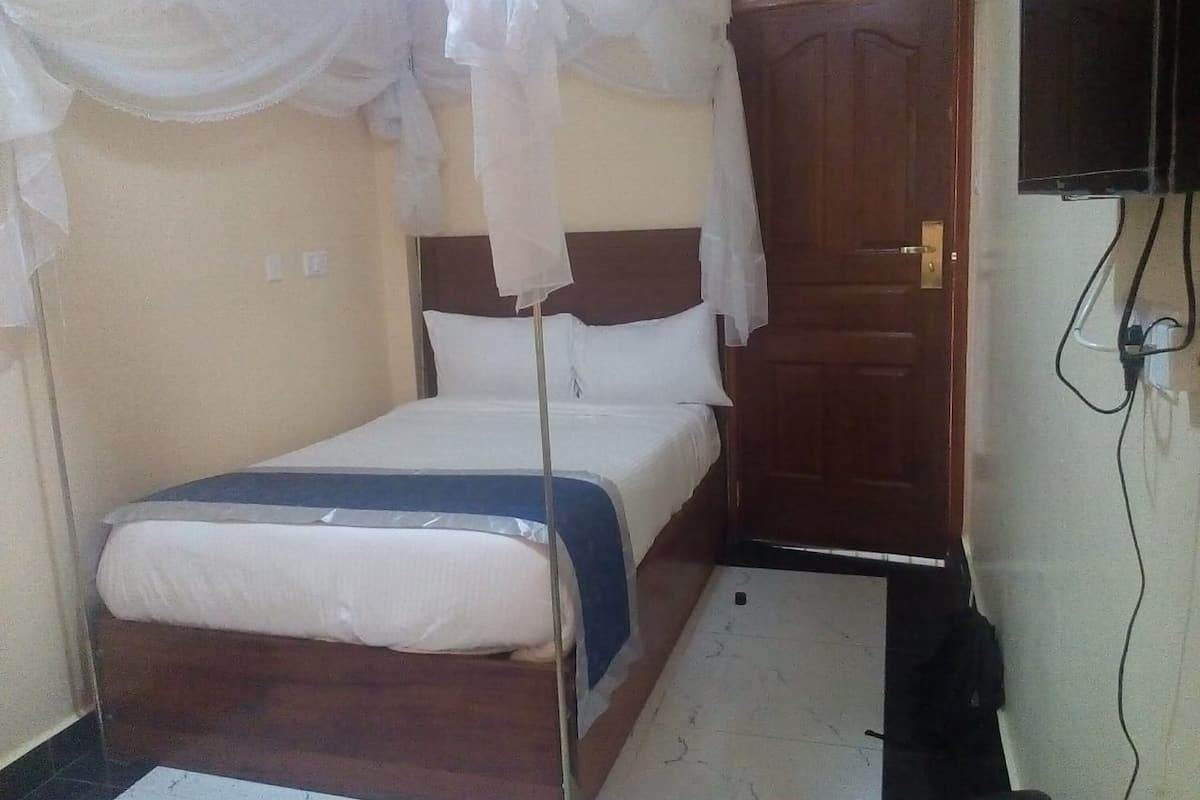 Una habitación pequeña con cama individual, cabecera de madera, mesita de noche, televisor montado en la pared y puerta con manija.
