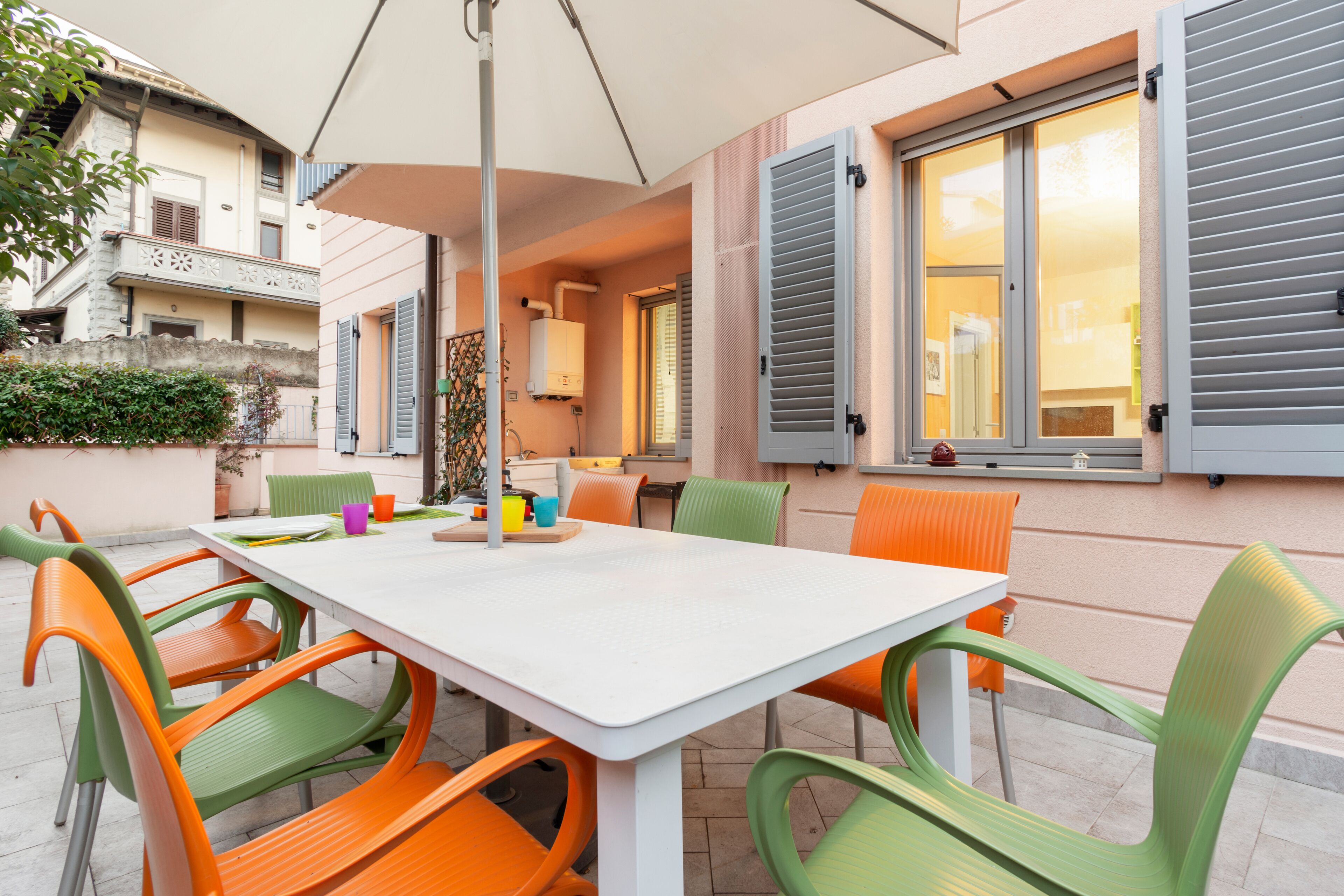 Un coin repas avec une table blanche et des chaises colorées, un grand parasol et une vue sur un bâtiment avec des volets.