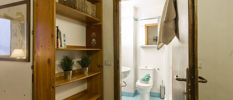 A wooden shelf unit with books and a small plant, leading to a bathroom with a toilet, sink, and tiled floor.