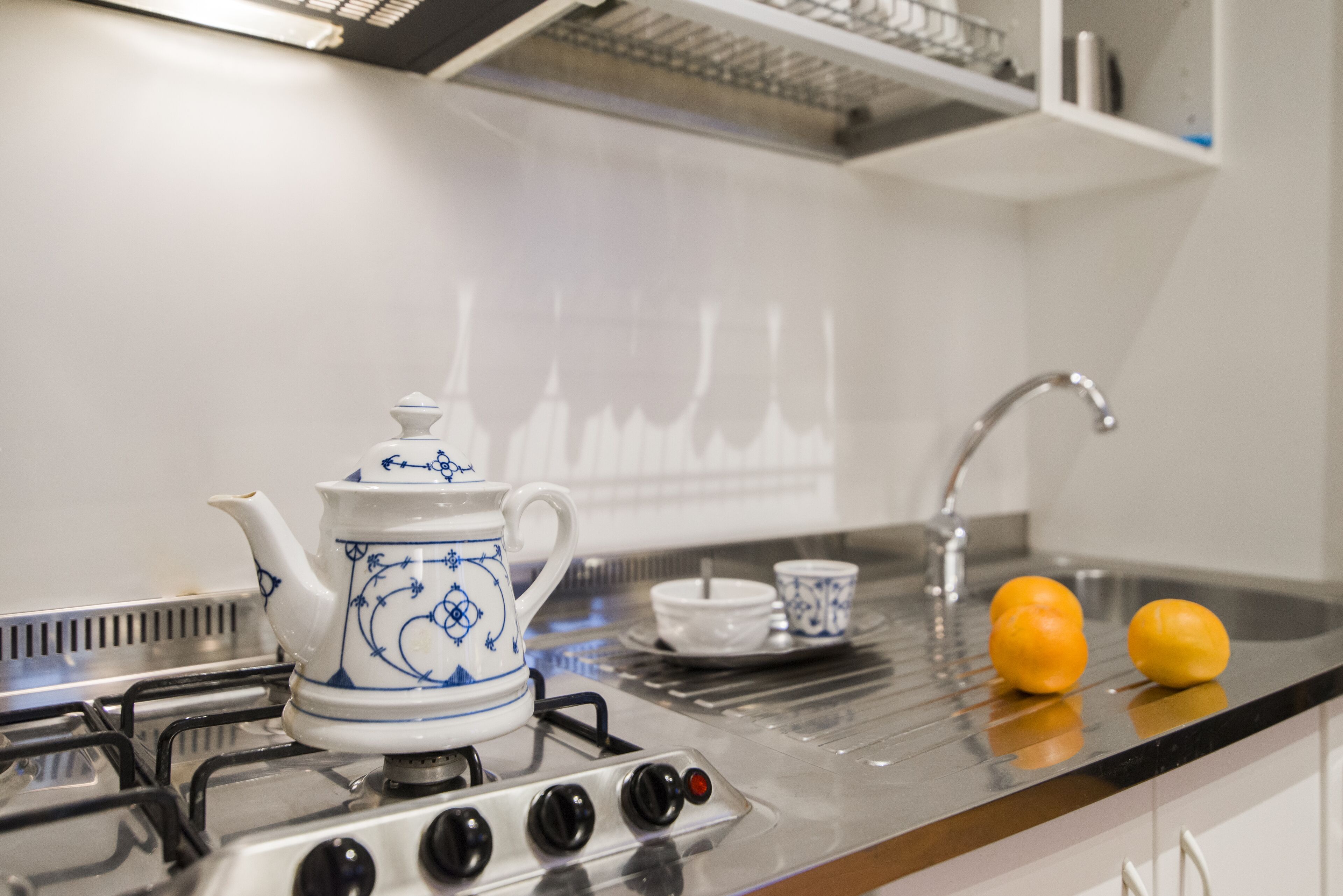 A kitchen with a gas stove, a white teapot with blue designs, a sink, and two oranges on the counter.