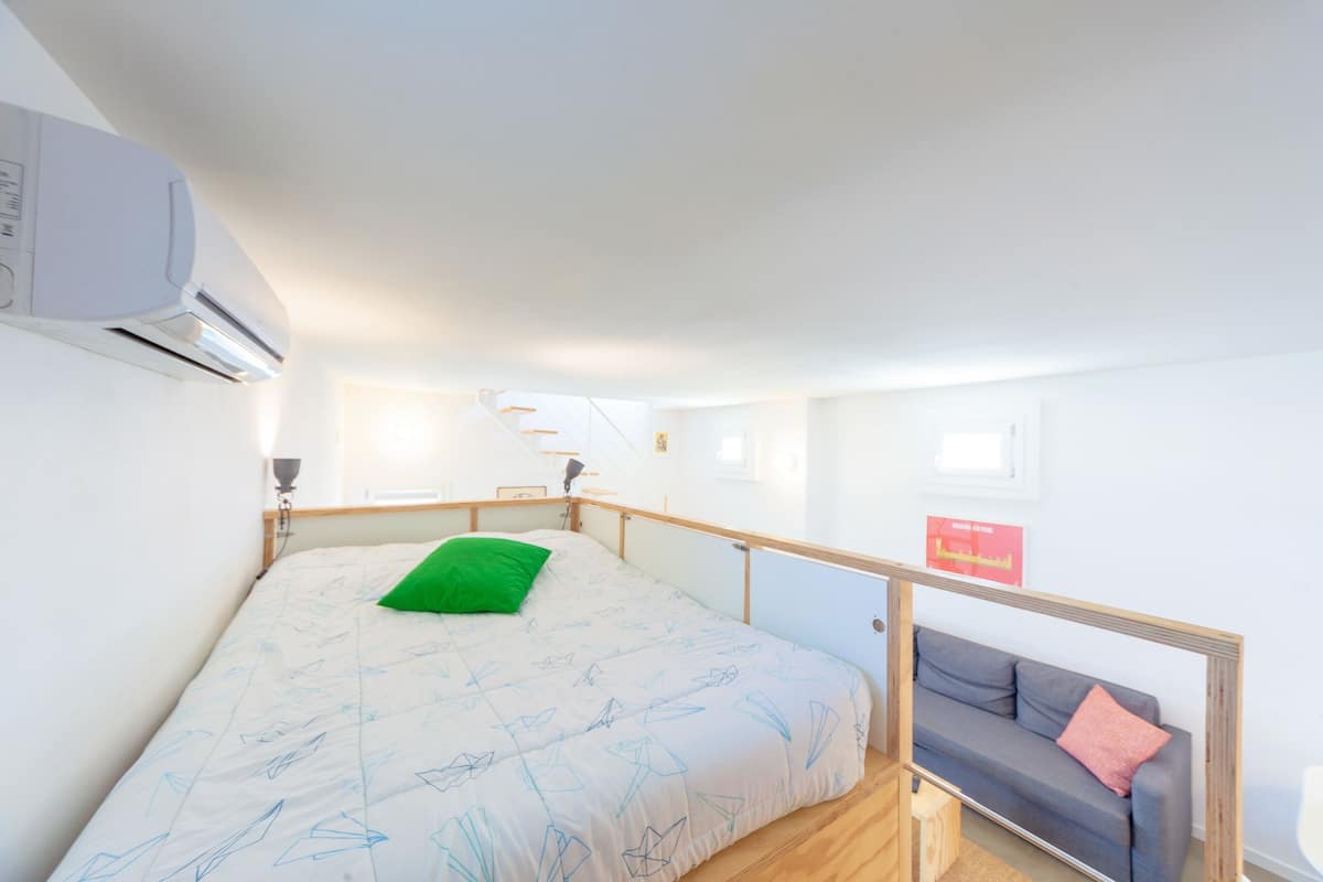Un dormitorio con una cama, un almohadón verde, un sofá gris y un aire acondicionado blanco.
