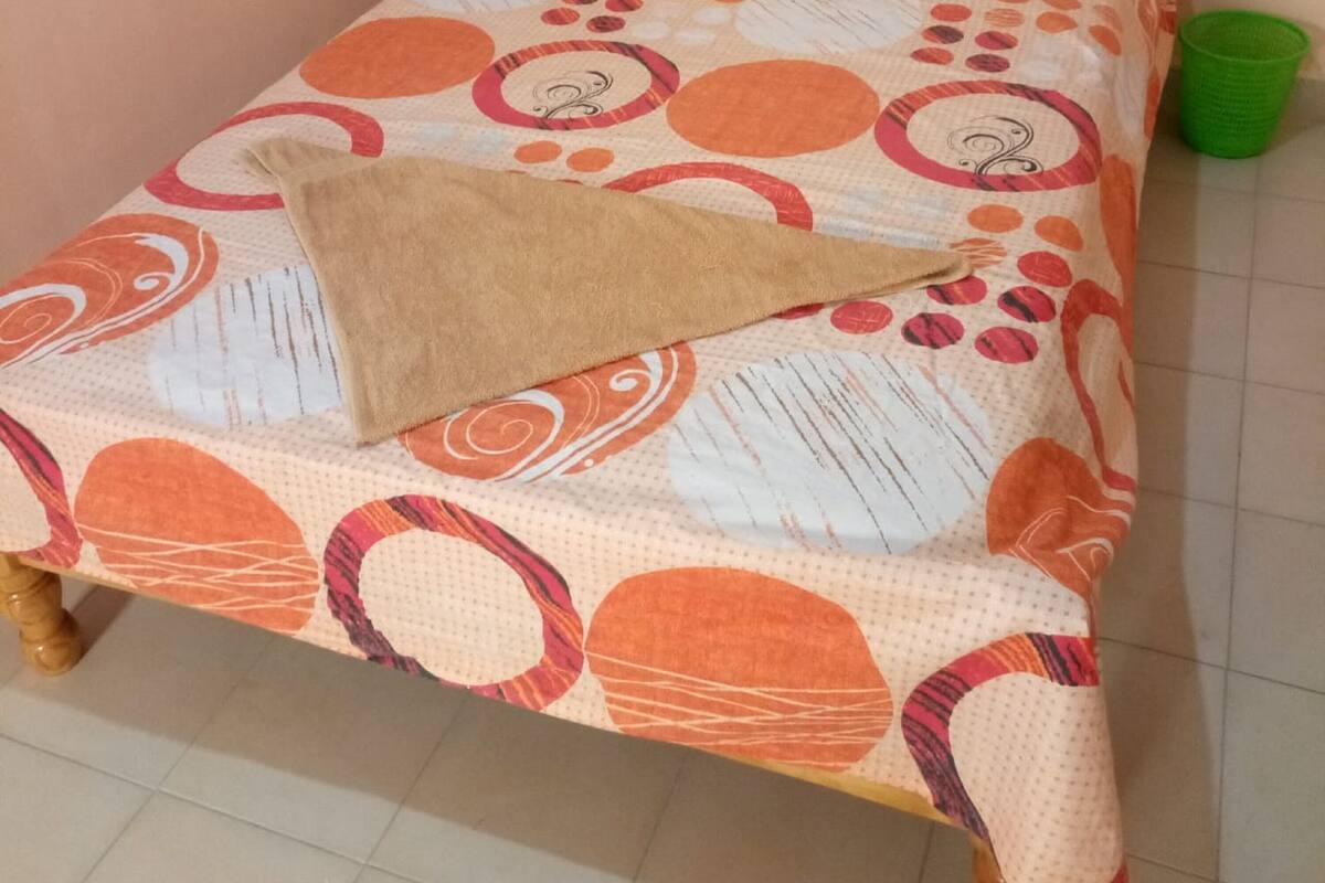 A single bed with a patterned bedspread, a pillow, and a green plastic container on the floor.