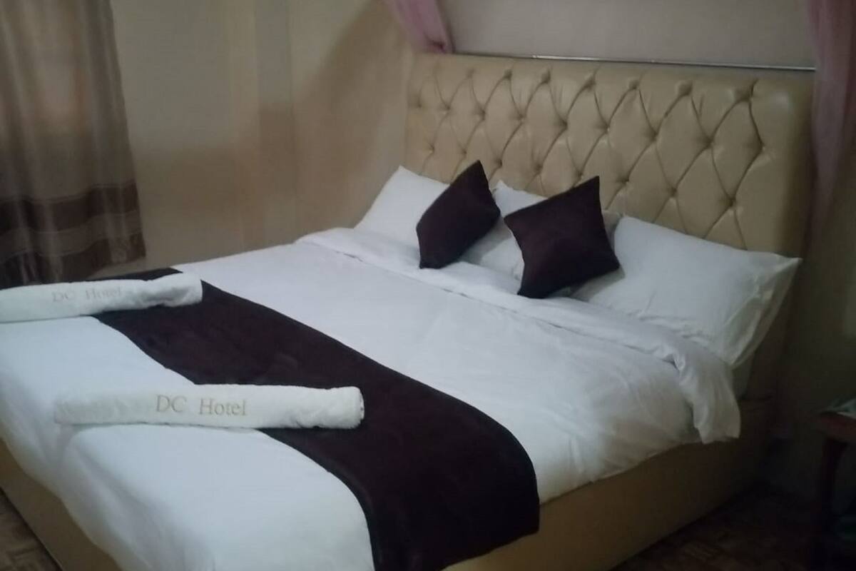 A neatly made bed with a white duvet, dark brown throw, and matching pillows, along with a set of white towels with "DC Hotel" branding.