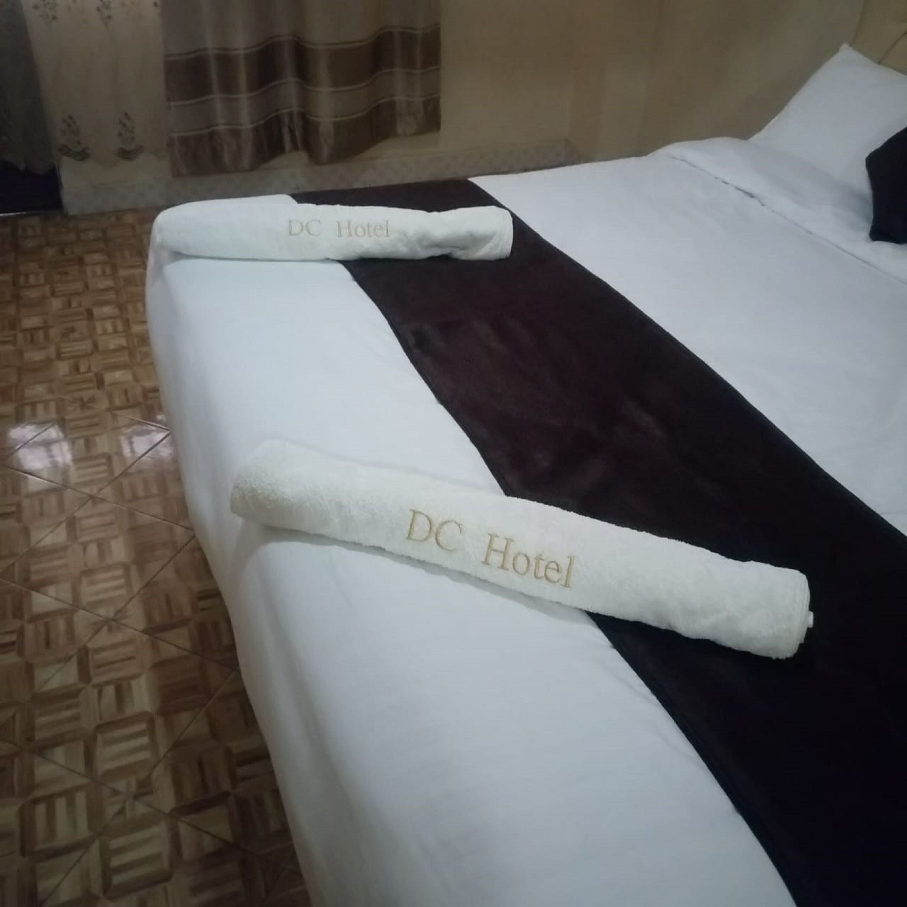 A hotel room with a neatly made bed, a dark brown bedspread, and a white towel with "DC Hotel" embroidered on it.