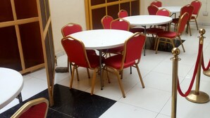 Une salle à manger d’hôtel avec des tables rondes et des chaises rouges, un sol carrelé en noir et blanc, et des stanchions dorées surmontées de cordes rouges.