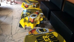 A bar area with stools covered in yellow and black 'Tusker' branded bags, a counter with bottles, and a sign advertising 'County Imewali'.