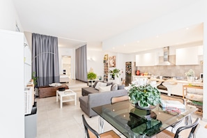 A modern living room with a glass dining table, grey sofa, and a kitchen area with white cabinets and appliances. - Luminoso Nuovalucello Apart- Giardino e Parcheggio privato (Catania)