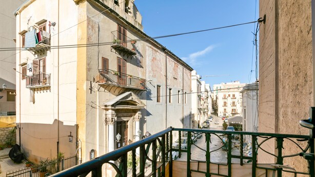 Eine enge Straße mit Gebäuden an beiden Seiten, ein Balkon mit grüner Reling und ein klarer, blauer Himmel.