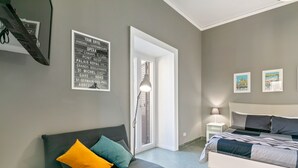Ein modernes Schlafzimmer mit einer grauen Couch, einem Bett mit grauer Bettwäsche, einem weißen Nachttisch und einer Lampe. An der Wand hängen gerahmte Bilder und ein Schild mit Text.