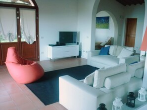 A modern living room with a white sofa, a red bean bag chair, a flat-screen TV on a white cabinet, and a painting on the wall.
