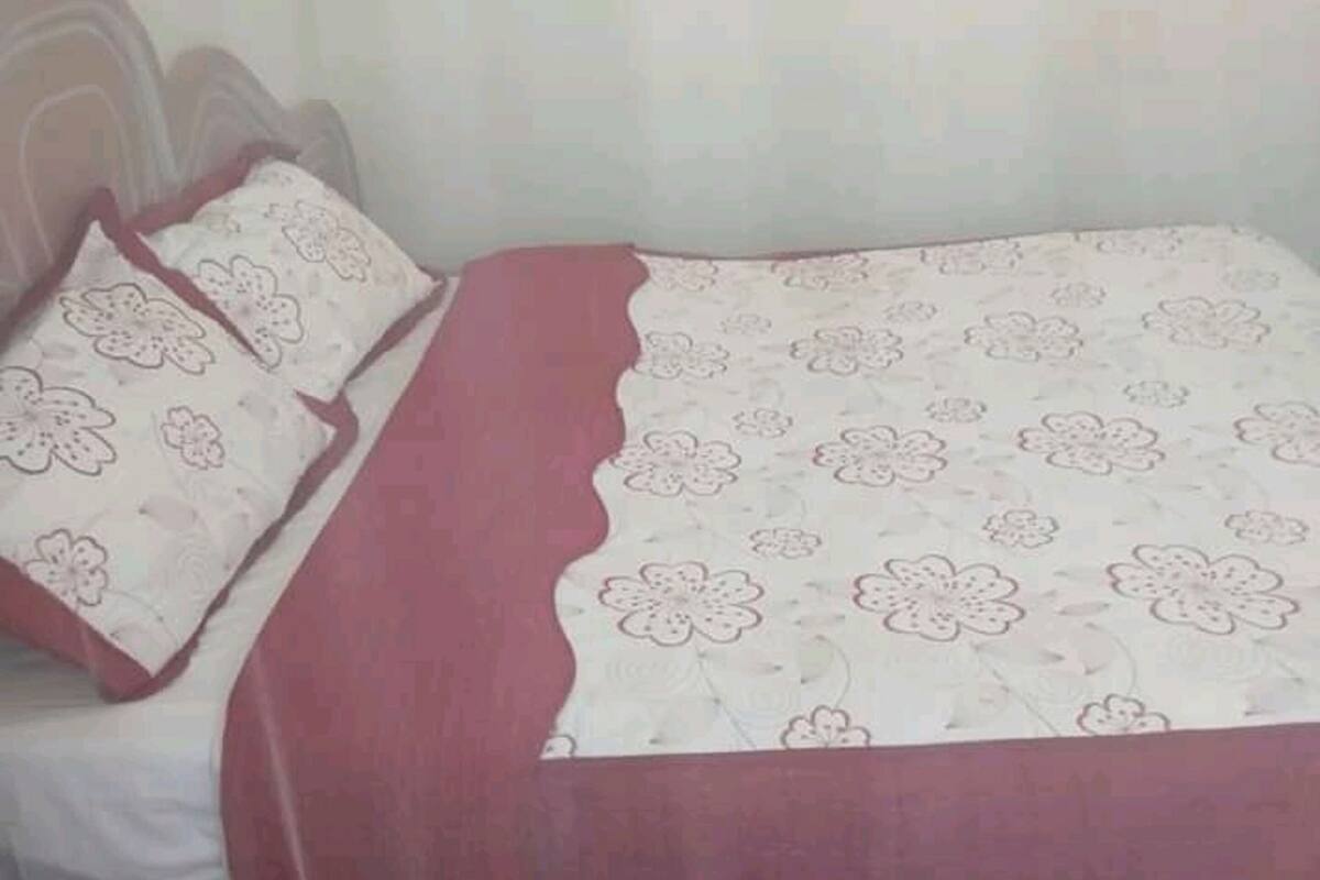 A bedroom with a bed covered in a floral-patterned bedspread, two matching pillows, and a sheer pink curtain.
