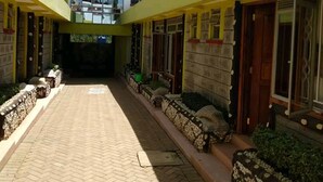 A courtyard with a paved walkway, yellow walls, and a building with a red roof in the background. - Kape Samaritan Guest House (Makutano)