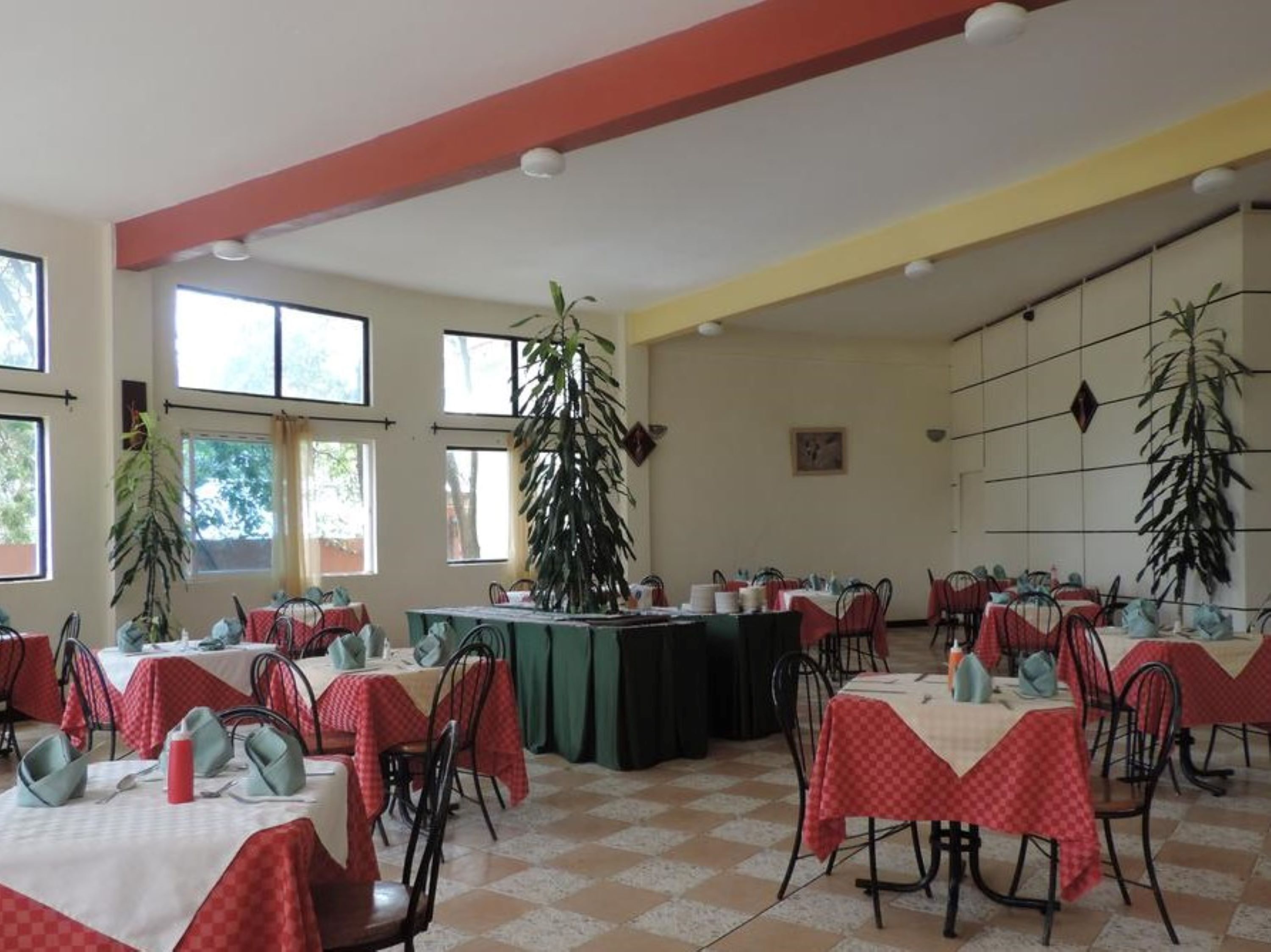 Uma sala de jantar com mesas preparadas para o serviço, grandes janelas e plantas em vasos.