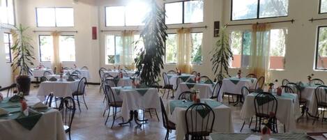 Un'ampia sala da pranzo con diversi tavoli e sedie, grandi finestre e piante in vaso.