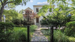 Une maison blanche dotée d’un porche à l’avant, entourée d’une végétation luxuriante et accessible par un chemin menant à l’entrée.