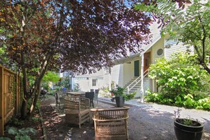 A covered outdoor seating area with wicker furniture, a potted plant, and a gravel path leading to a house.