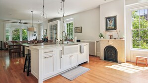Une cuisine spacieuse dotée d’un grand îlot central, de planchers en bois, de luminaires suspendus, d’une cheminée et d’une vue sur la verdure grâce à de grandes fenêtres.