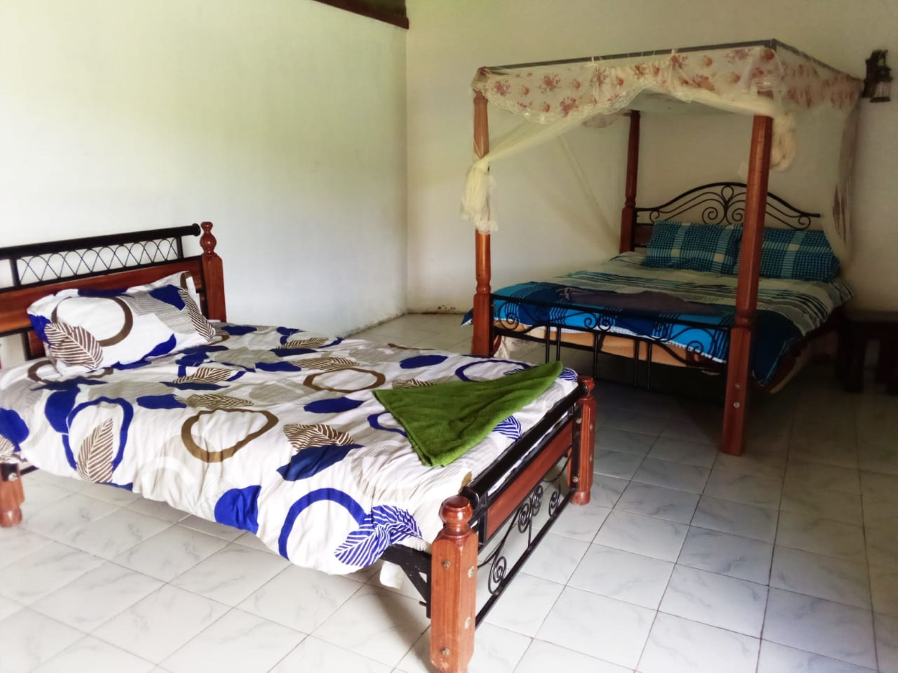 A room with a single bed with a floral canopy, another bed with a patterned bedspread, and a tiled floor.