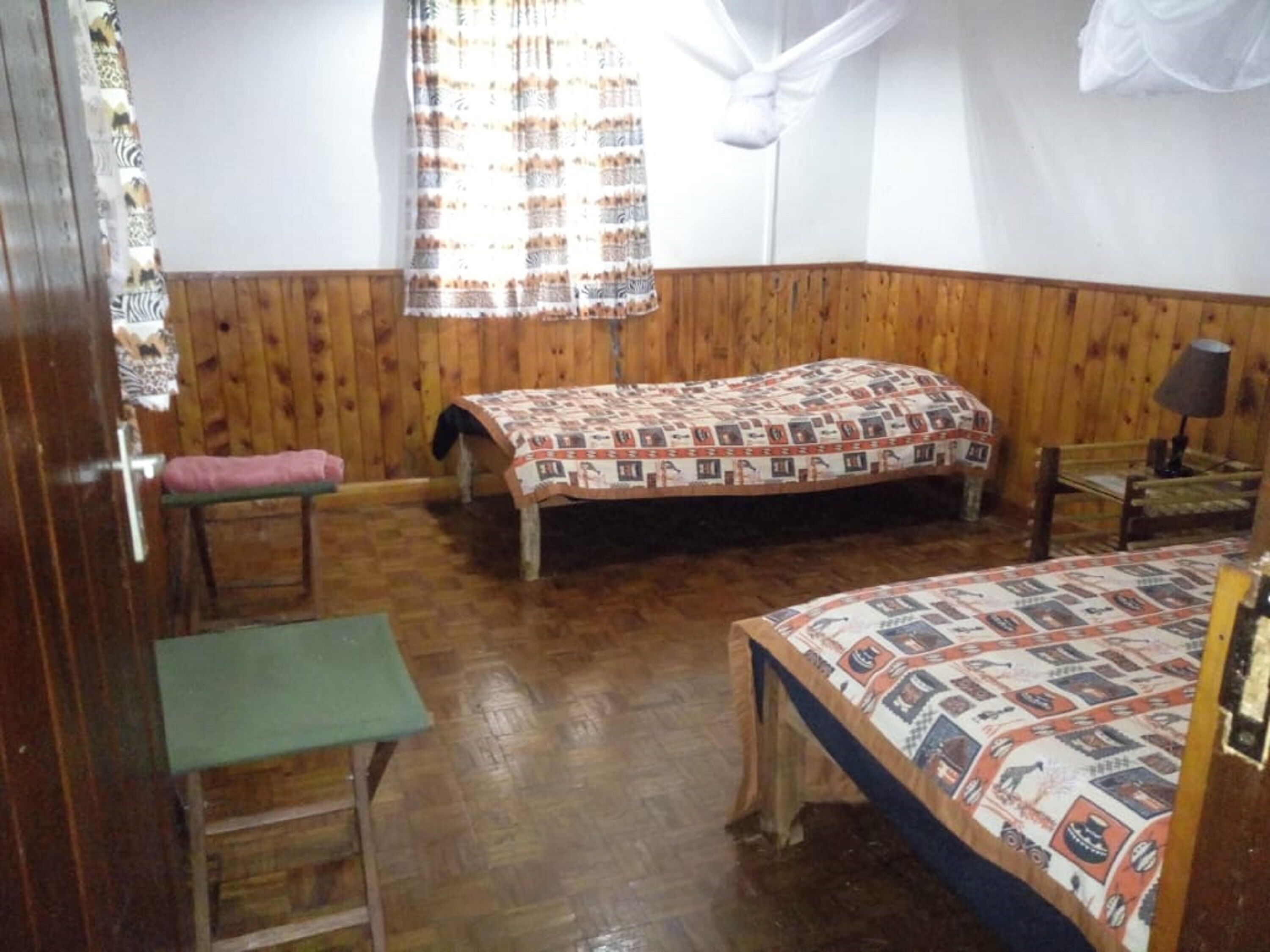 A room with wooden walls and floors, a bed with a patterned bedspread, a bench, a small table, and a window with a white curtain.