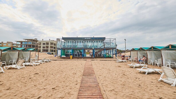 Un edificio frente a la playa con vista despejada, un paseo de madera que lo conduce y varias sillas de playa dispuestas a ambos lados.
