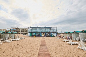 Un bâtiment situé en bord de mer, offrant une vue dégagée sur la plage, avec une promenade en bois menant à la plage et plusieurs chaises de plage disposées de part et d’autre.