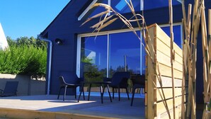 Una casa moderna con facciata blu, ampie finestre e un terrazzo in legno con mobili da esterno.