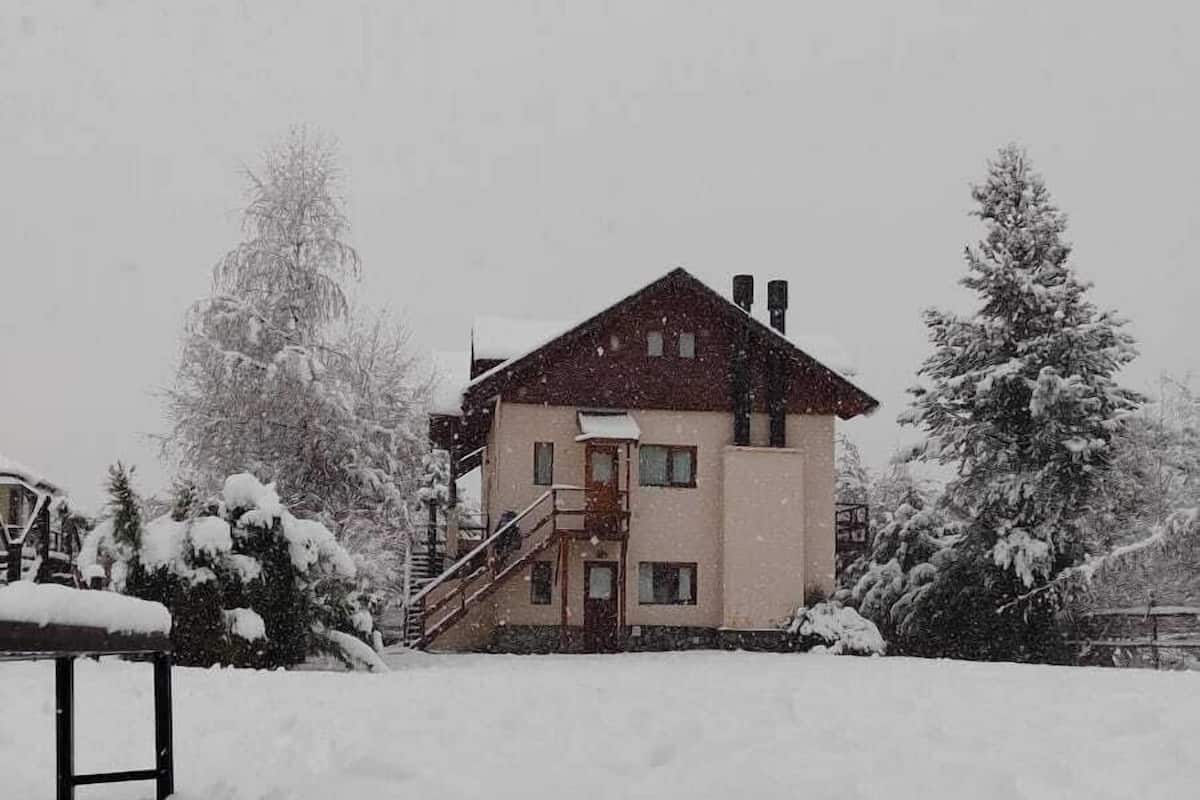 A two-story house with a red roof and beige walls, surrounded by snow-covered trees.