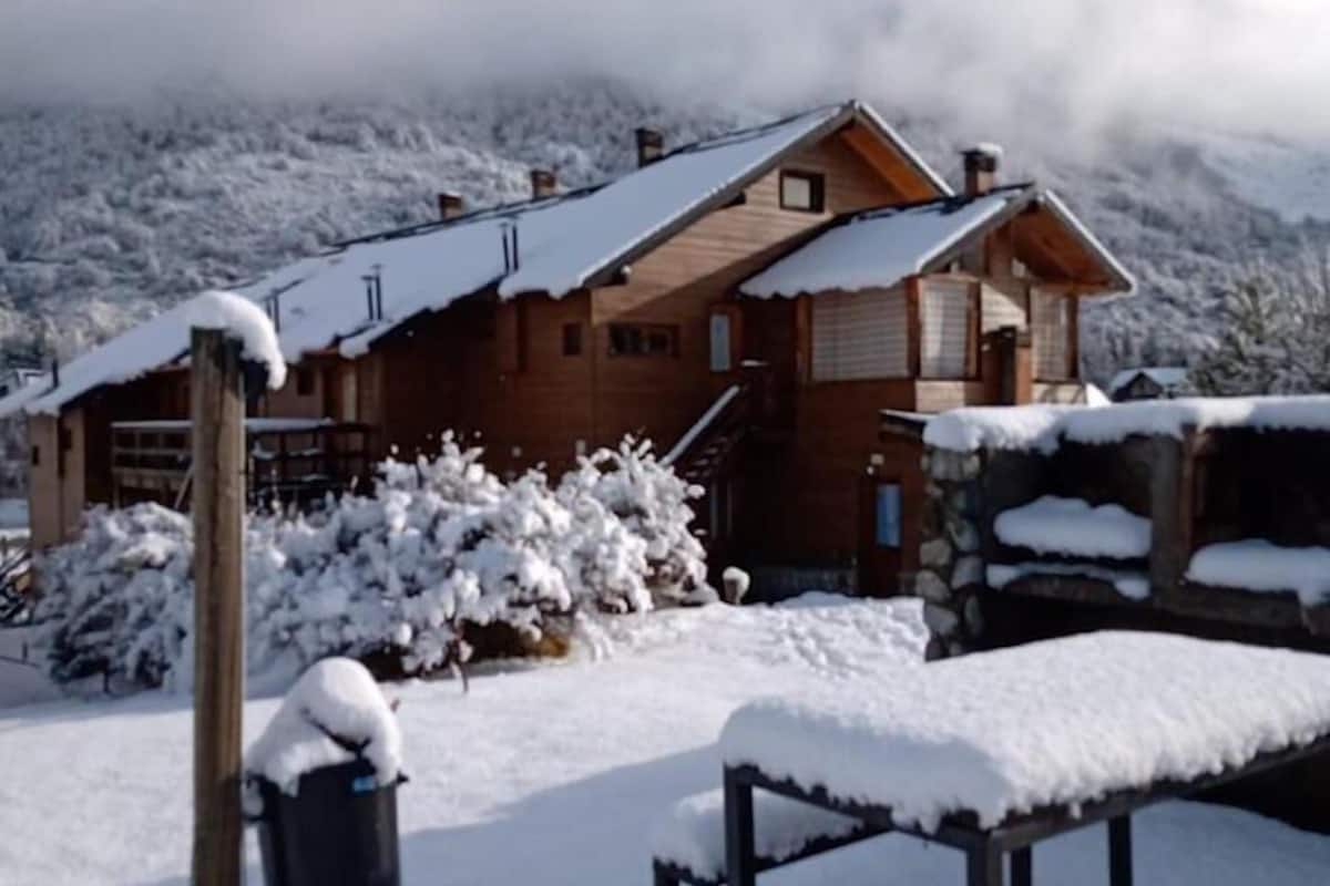 A wooden chalet with a snow-covered roof, surrounded by snow-covered trees and a mountainous backdrop.