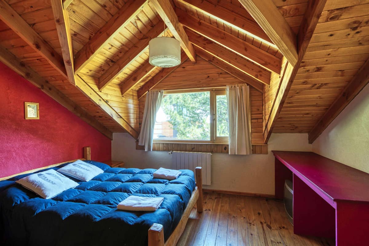 A bedroom with a sloped wooden ceiling, a bed with blue bedding, a window with curtains, and a red desk.