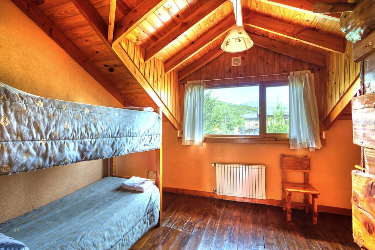 A room with a sloped ceiling, a window with curtains, a radiator, a wooden chair, and a bunk bed.