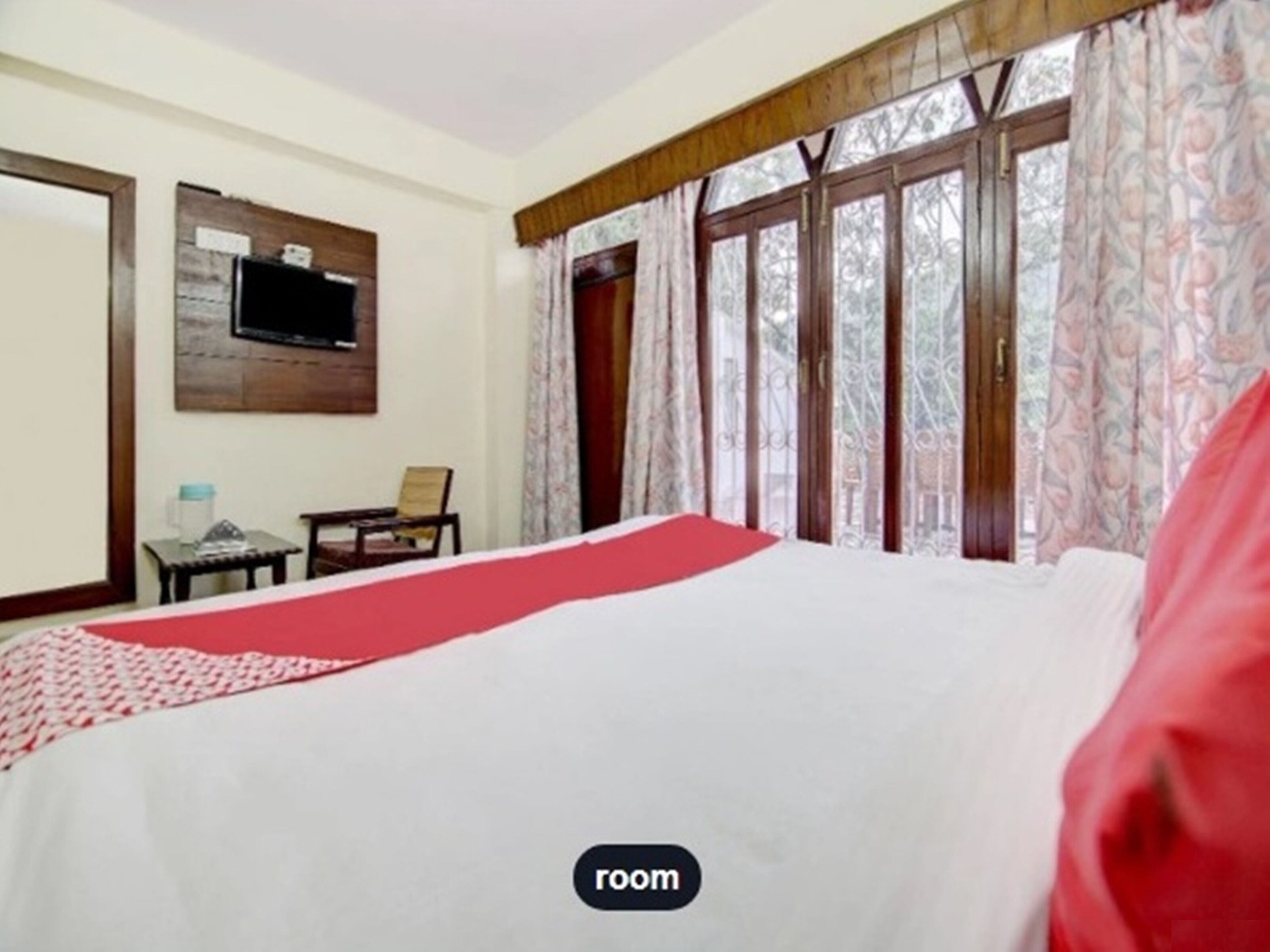 A hotel room with a bed, a television mounted on the wall, a small table with a chair, and a window with curtains.