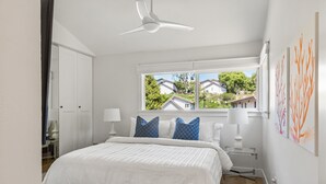Une chambre à coucher avec un lit, des murs blancs, un ventilateur de plafond, une fenêtre donnant sur des maisons et des œuvres d’art accrochées au mur.