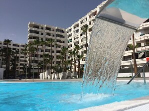 Une piscine agrémentée d’un élément décoratif aquatique, entourée de palmiers et de bâtiments à plusieurs étages.