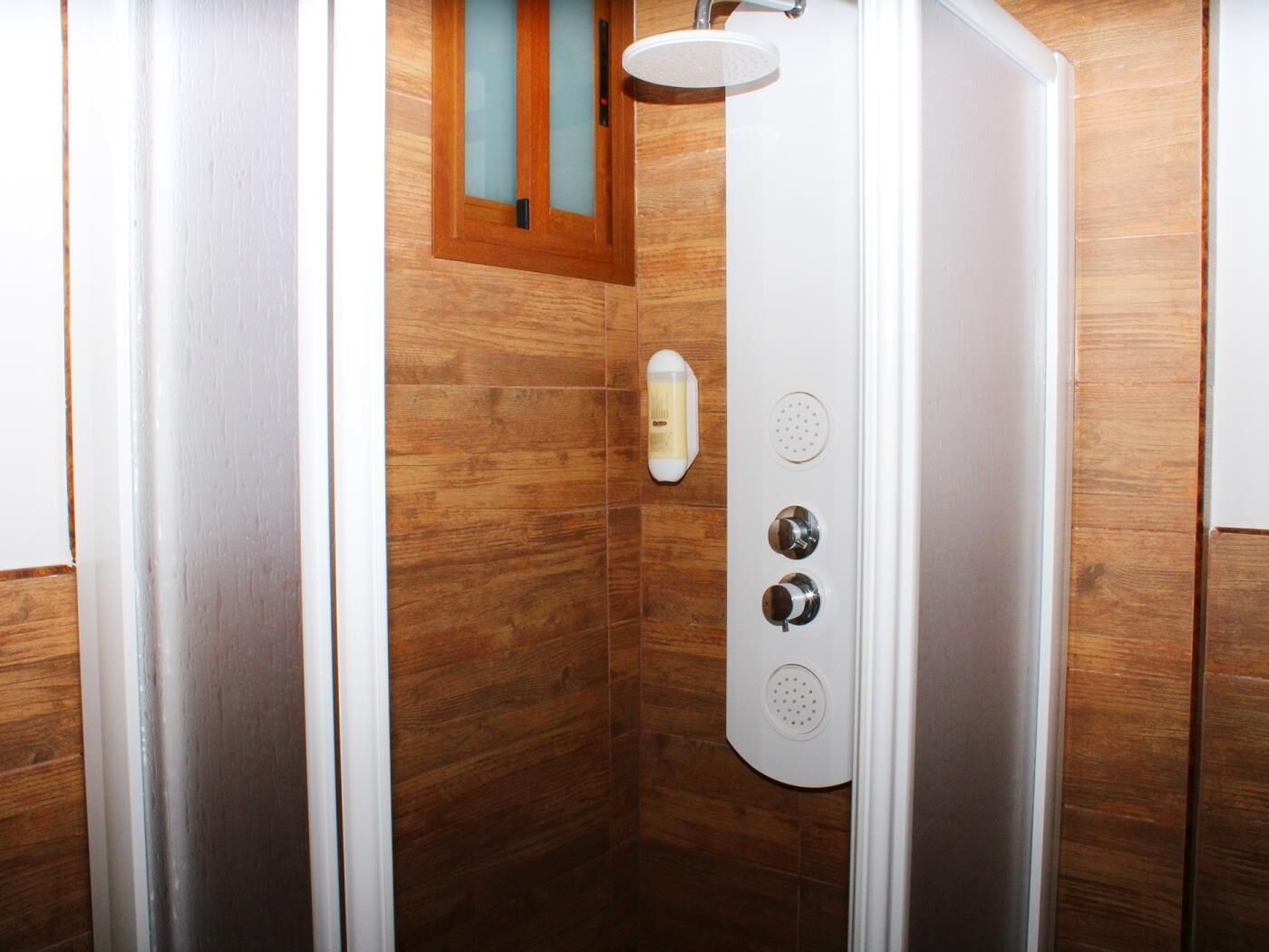 A shower with a control panel, a soap dispenser, and a window above it.