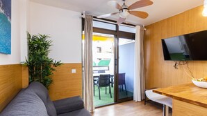 A modern room with a grey sofa, wooden accents, a ceiling fan, a TV, and a view through a glass door to an outdoor area.