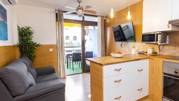 A compact living space with a kitchenette, a grey sofa, a ceiling fan, and a TV mounted above the counter.